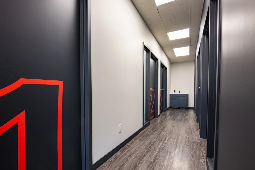 Modern office hallway with dark gray doors featuring large red room numbers, wood-look flooring, white walls, and bright ceiling lights leading to a cabinet at the end of the corridor.