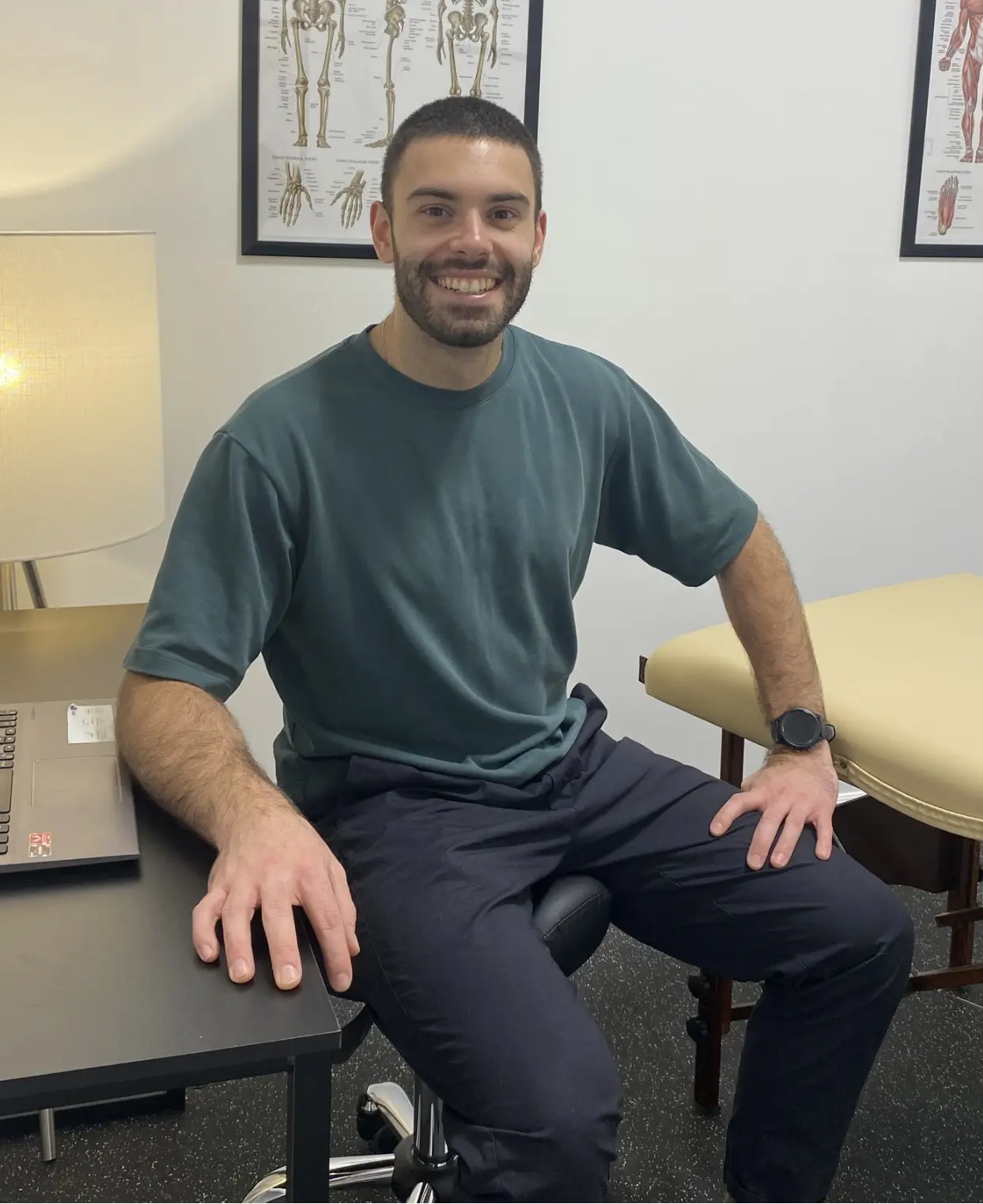 "Osteopath seated in a consultation room with an examination table and anatomical charts."